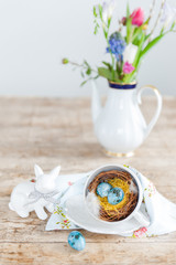 Bright easter eggs in a nest from a cup in the foreground. Porcelain hares and a vase with flowers in the background