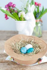 Rustic wicker hat with bright Easter eggs in the foreground and a bunny vase with flowers in the background