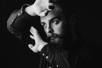Black and white portrait of a stylish man with a beard and stylish hairdo dressed in the black shirt holding his hands in front of his face on the black background