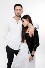 Beautiful brunette girl in a white tulle skirt in a black bra and black leather jacket with guy in trousers and white t-shirt are hugging on the white background in the studio