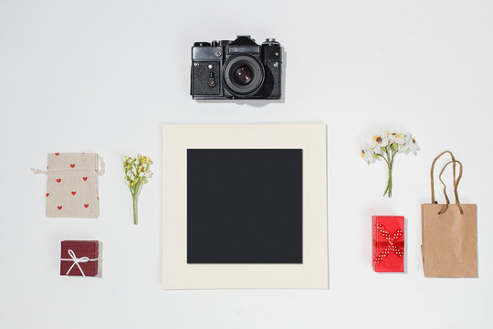 Composition with black photo frame, retro camera, gift boxes, craft bag, canvas bag with red heart shapes and spring field flower on white desk. Trendy flat lay mockup for designers, photographers