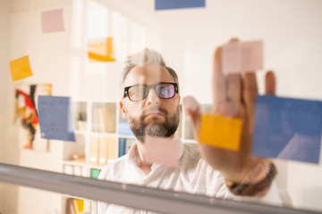 Serious introspective handsome businessman in eyeglasses joining stickers on glassy board while planning new project in office