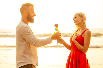 man in a white shirt giving roses to surprised wonder woman in a long red dress on the ocean beach. romantic date, wedding or valentines day concept by sea.loving couple celebrating March 8 Women Day
