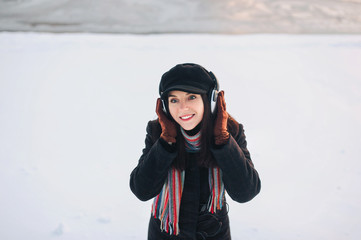 Fototapeta na wymiar A young beautiful girl in gray headphones in winter on a snow background. To cheer up the music. Playlist with your favorite songs. Spreading hands. Copy space.