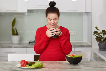 Expectant young mother has breakfast at kitchen, eats fresh vegetable salad and drinks tea, wears casual red sweater, has healthy eating, tastes aromatic beverage. Motherhood and eating concept