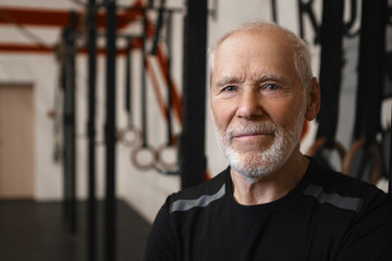 Close up shot of mature seventy year old Caucasina man with wrinkles, blue eyes and thick beard wearing stylish black t-shirt while exercising in gym alone, looking at camera with happy smile