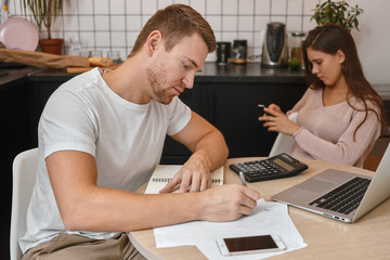 Fototapeta premium Portrait of handsome adult male studying at home, sitting at table in front of portable computer, making notes in copybook, having online marketing courses, cute woman using cell phone in background