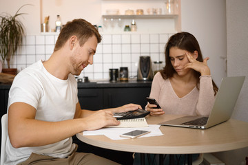 Obraz premium Stylish young serious man freelancer working from home using laptop, sitting at kitchen table, studying papers, reading them attentively, his cute wife scrolling social networks on mobile phone