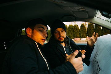 Two armed bandits sitting in a car planning their next robbery, while counting on a stopwatch the time they have for the robbery, showing off their guns and pointing at the blueprint.