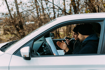 Naklejka premium Two armed bandits sitting in a car planning their next robbery, while counting on a stopwatch the time they have for the robbery, showing off their guns and pointing at the blueprint.