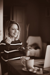 Young lady in sweater sitting at the tablebreakfast time with cup of coffee and cake and looking in to notebook computer. Image in sepia color style