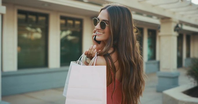 Beautiful brunette woman shopping walking through the mall glances back at the camera with phone and shopping bag in hands