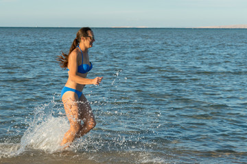 A sexy young brunette woman or girl wearing a bikini running through the surf on a deserted tropical beach with a blue sky. Young woman running by the sea
