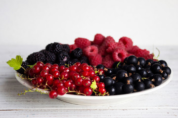 Black and red currants, black and red raspberries on a plate with a leaf of raspberry, white