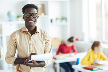 Succesful young school teacher in shirt and eyeglasses looking at you while making notes