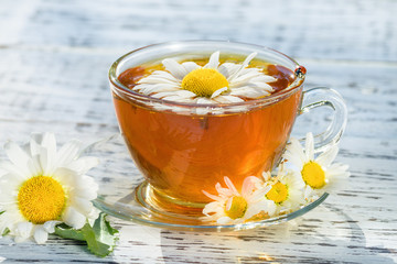 Cup of tea with chamomile, on which there is a ladybug, close-up, illuminated by sunlight, on a white wooden table.