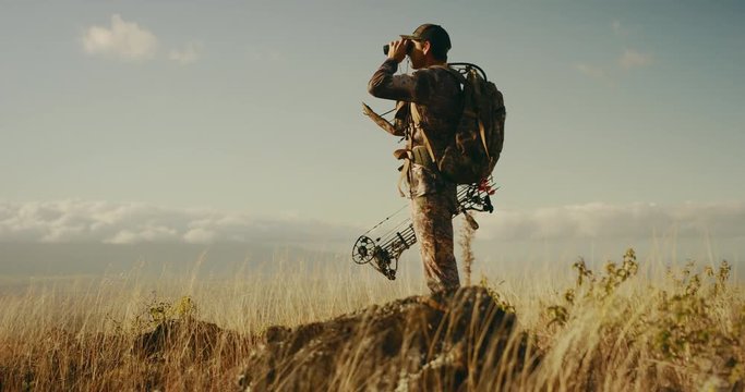 Orbit shot of majestic bowhunter on grassy knoll with his binoculars raised