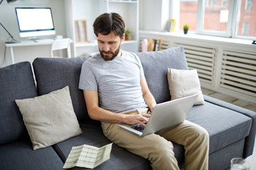 Young accountant looking at financial data on paper wnd entering it in laptop while working on sofa in home office