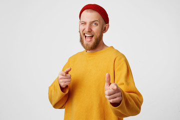 You're chosen bro. Yes, we did this! Indoor shot of cheerful bearded man in red hat and casual sweater indicates happily at you, chooses to compete, has positive expression, isolated on white wall