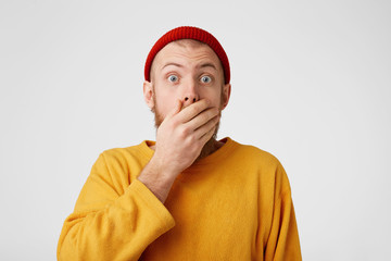 Surprised young man covers his mouth with palm, stares at camera with bugged eyes, feels shocked to hear sudden news, isolated over white background