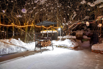 BBQ near the home in the winter. Night, garlands are burning, it is snowing. Empty playground. They lay harvested firewood under the grill