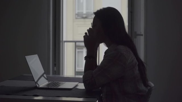 Silhouette of tired young woman with headache working with laptop. Side view of tired businesswoman working on project at home. Overwork concept
