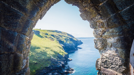View through the ruins of Tintagel castle of British west landscape coastline, shore for summer holidays. Cornish summer with green rocky cliffs in Tintagel, Cornwall, UK.  