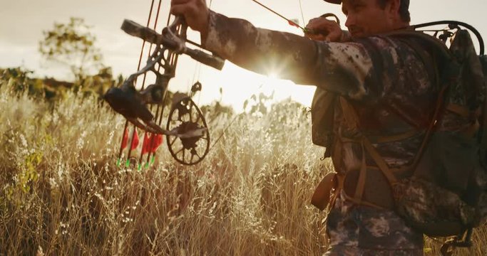 Camera tilts up from the grass to bowhunter as he draws back his bow, preparing to shoot