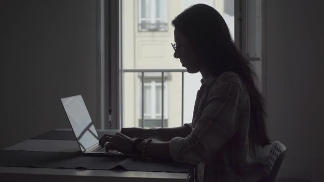 Focused hindu freelancer in eyeglasses working with laptop at home. Side view of serious young businesswoman typing on laptop while sitting near window. Technology concept