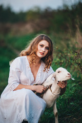 .A girl in a white dress walks with a goat in nature.