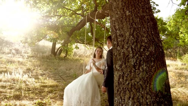 Groom shakes his bride on swing and smiles with loving look. Woman and man in romantic meeting are happy in highlights of sunset.