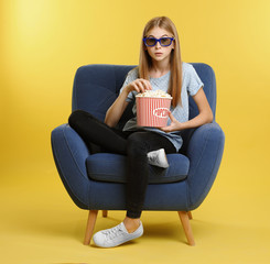Emotional teenage girl with 3D glasses and popcorn sitting in armchair during cinema show on color background