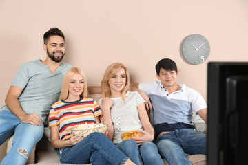 Young friends with snacks watching TV on sofa at home