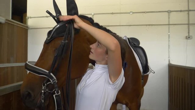 SLOW MOTION, CLOSE UP: Young woman tacking up and putting a bridle on her beautiful brown horse before a dressage competition. Happy girl sliding a bit into her stallion's mouth while tacking him up.