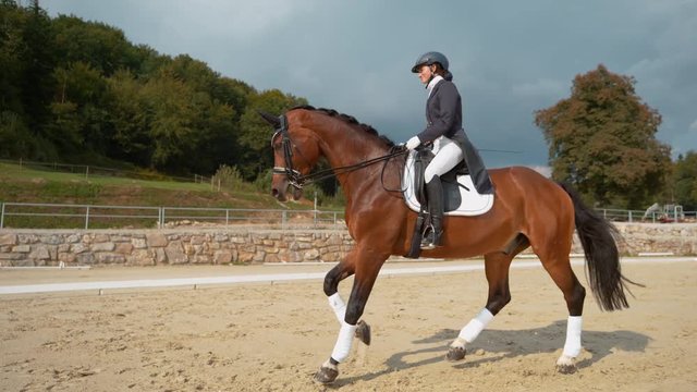 SUPER SLOW MOTION: Young woman in a dark blue frock having fun galloping on her stunning chestnut stallion. Female English riding training her horse and galloping around the sandy arena in countryside