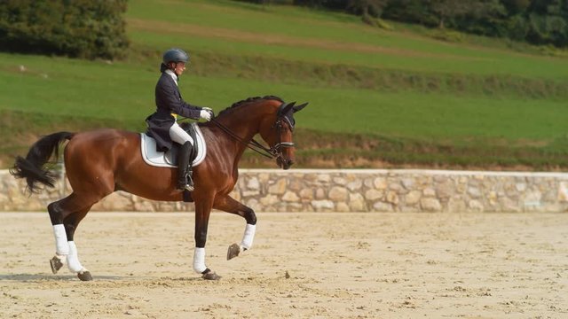 SUPER SLOW MOTION: English rider on horseback riding trot around the sandy arena. Elegantly dressed woman riding her stallion around the paddock during a dressage competition. Girl training her horse.