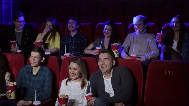 A group of young people sit in a cinema with popcorn and drinks and watch a funny comedy film, they laugh. Young friends stylish girls and boys watching new movie at cinema, spending time together.
