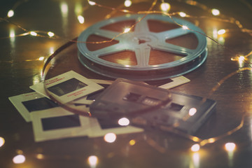 Photo slides, cassette audio tape and a 8mm or super 8 vintage film reel on a wood table with soft lights.