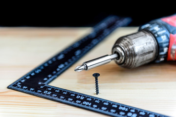 Electrical screwdriver, screw and measuring square on wooden board on black background.