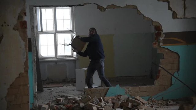 Depressed and mad man throws a chair through a room in an old abandoned house