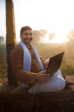 Smiling farmer using laptop while sitting near his agriculture field
