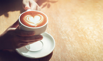 Close up girl hands holding latte art coffee in cup with white heart cream on top on wooden table. concept valentine coffee
