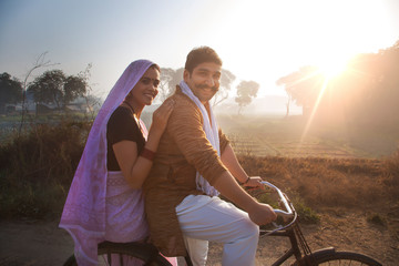 Side view of a happy rural couple in traditional dress sitting on bicycle in a village with agricultural fields in the background.	