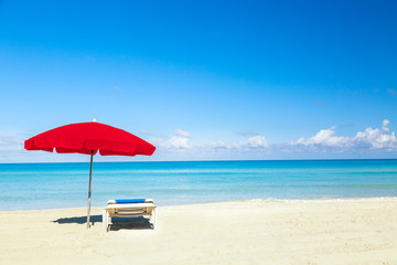 Fototapeta na wymiar A sun lounger under red umbrella on the sandy beach by the sea and sky. Vacation background. Idyllic beach landscape.