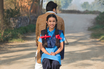 Smiling young school girl riding pillion on bicycle facing backward going along with her father in village.