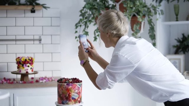 Confectionery blog. Woman in white shirt photographing cake decorated with flowers. Confectioner decorated the cake and then takes pictures on the smartphone. Side view