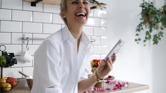 Cheerful, positive woman in white shirt standing on her white modern kitchen with smartphone in her hands, having fun, laughing. Confectioner decorated the cakes with flowers