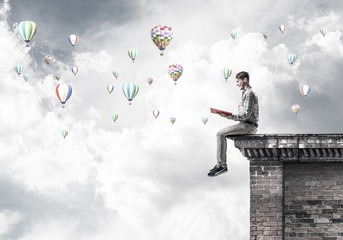 Man on roof edge reading book and aerostats flying in sky