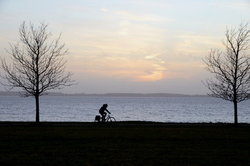Silhouette of a person on a bicycle in evening light, with the sea as background.