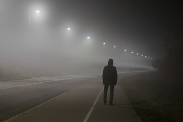Young man alone slowly walking under white street lights in night. Dark time. Peaceful atmosphere in mist. Foggy air. Back view.  - Powered by Adobe